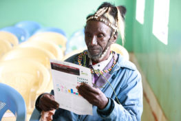 Village elder wearing a headdress reading the Itare-Chemosit Sub-Catchment Management Plan (SCMP) for forestry and water governance in Kenya Rau Valley