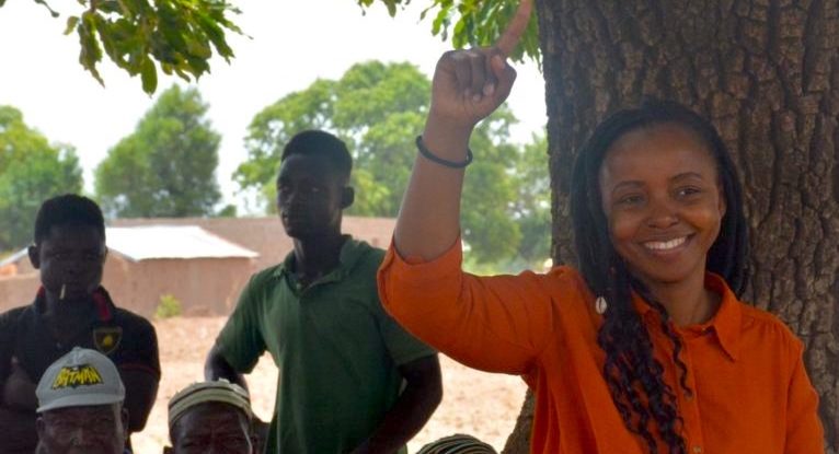 Woman stands beside tree, speaking with farmers