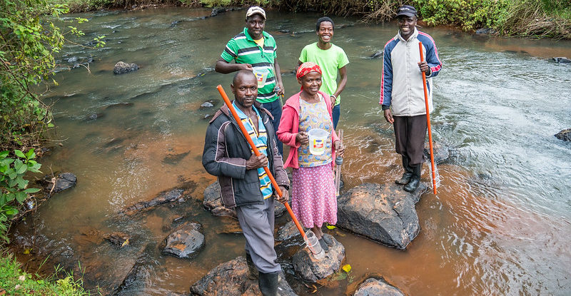 A group of five people stand on rocks in a river, some of them holding sticks with plastic water bottles attached to take samples