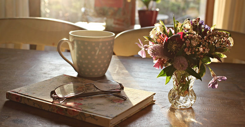 Tea cup, book, glasses and a small vase of flowers on a table